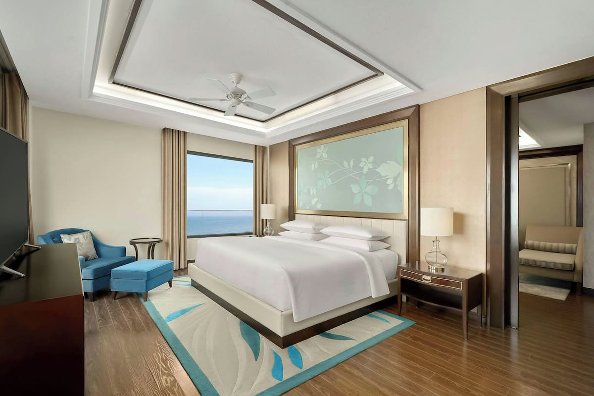 A hotel room with a bed, a desk, a chair, a TV, and a view of the ocean.