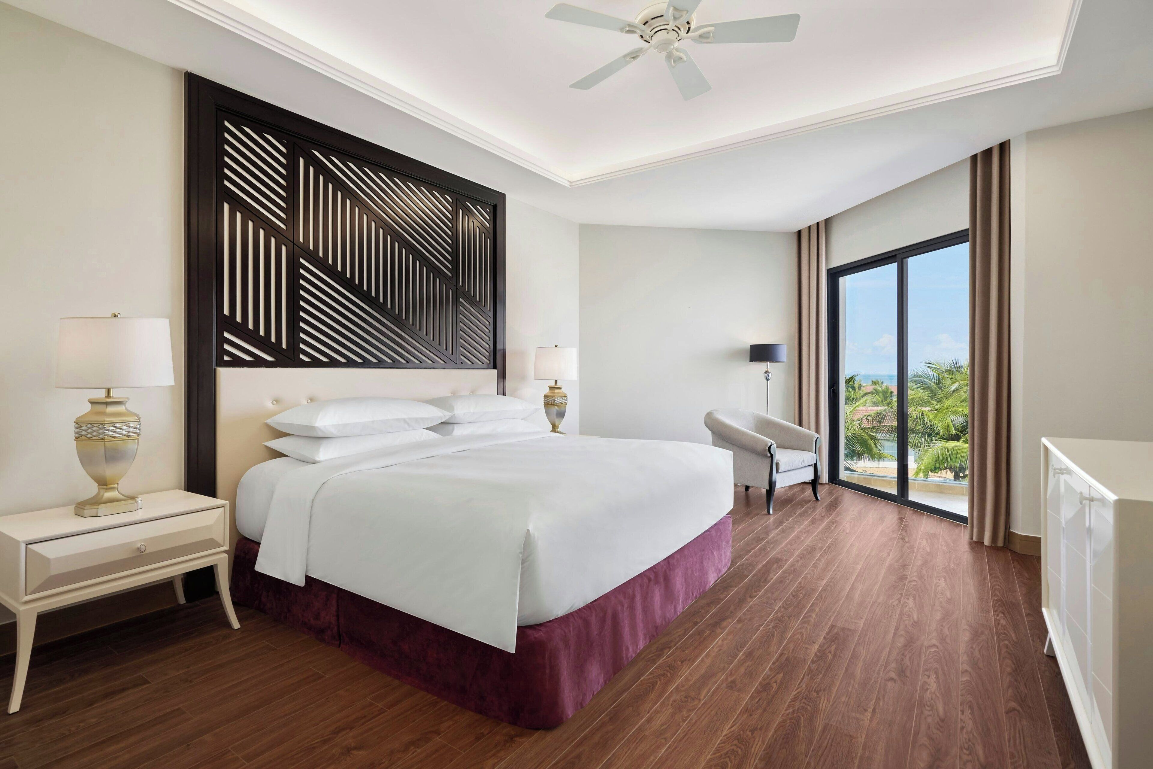 A hotel room with a large bed, a desk, a chair, a ceiling fan, and a view of the outdoors.