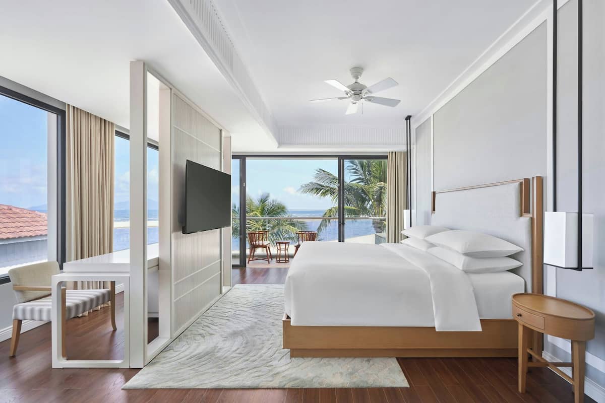A modern bedroom with a large bed, a TV, a balcony with a view of the ocean, and a ceiling fan.