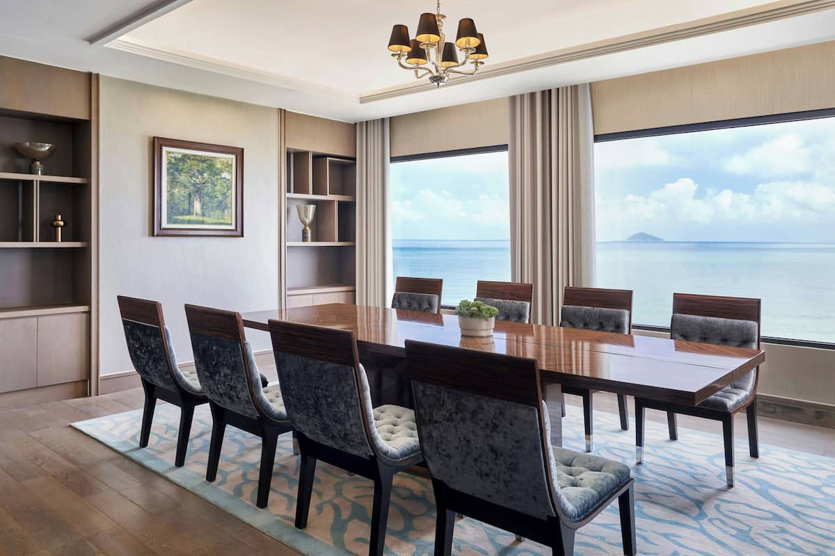 A dining area with a large table, six chairs, a chandelier, and a view of the sea.