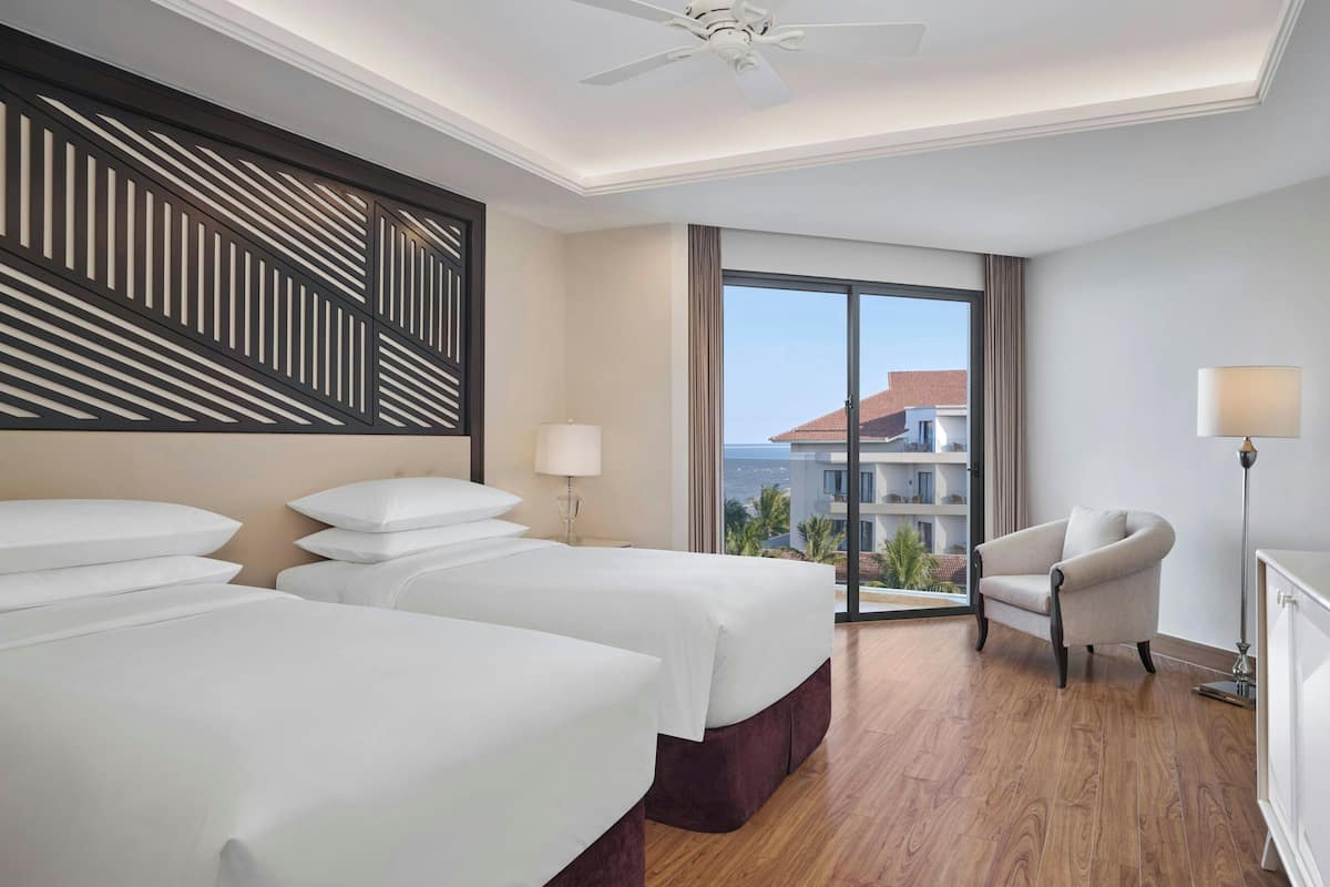 A hotel room with two beds, a chair, a lamp, and a view of the city.