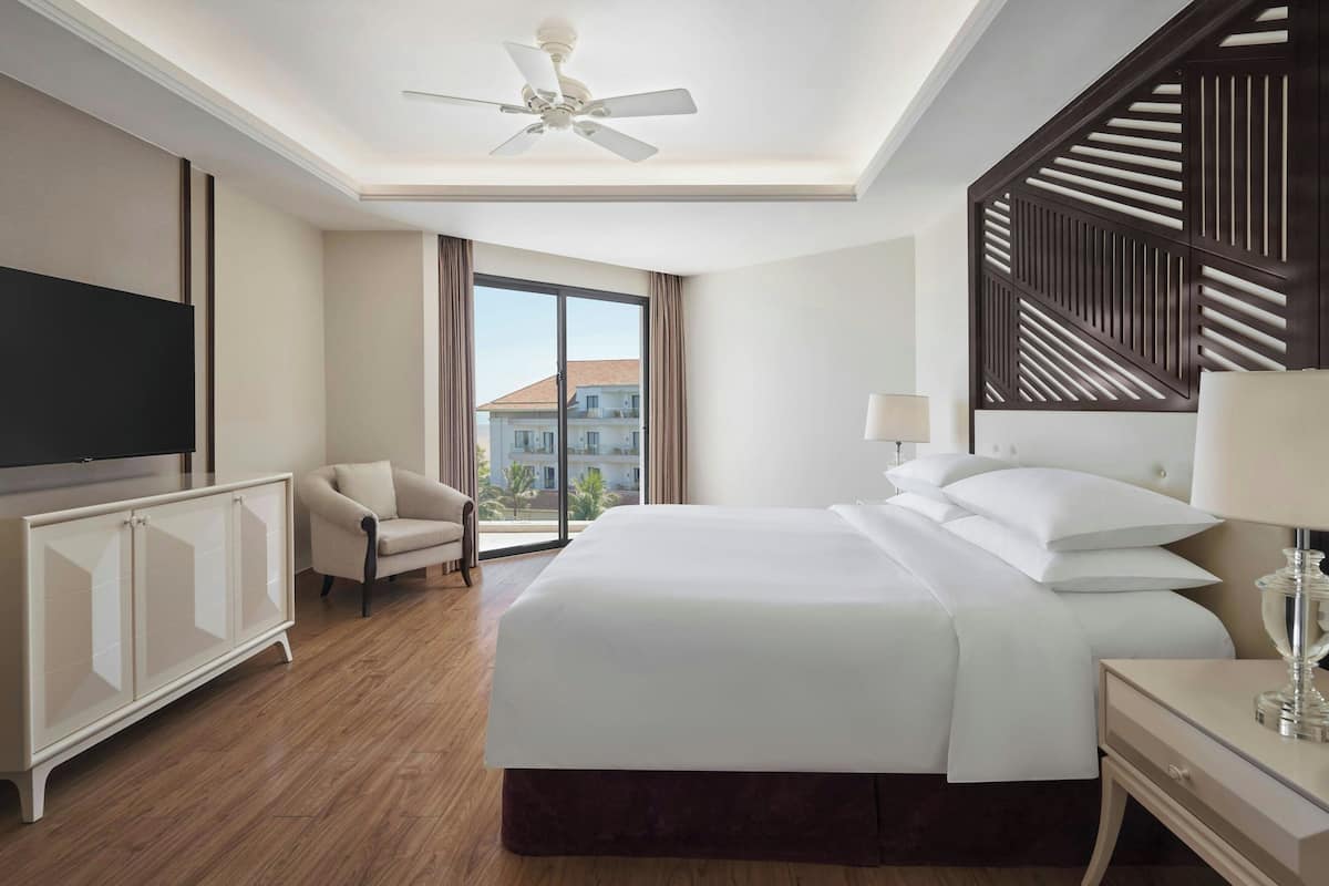 A hotel room with a large bed, a TV, a chair, a desk, and a view of buildings outside.