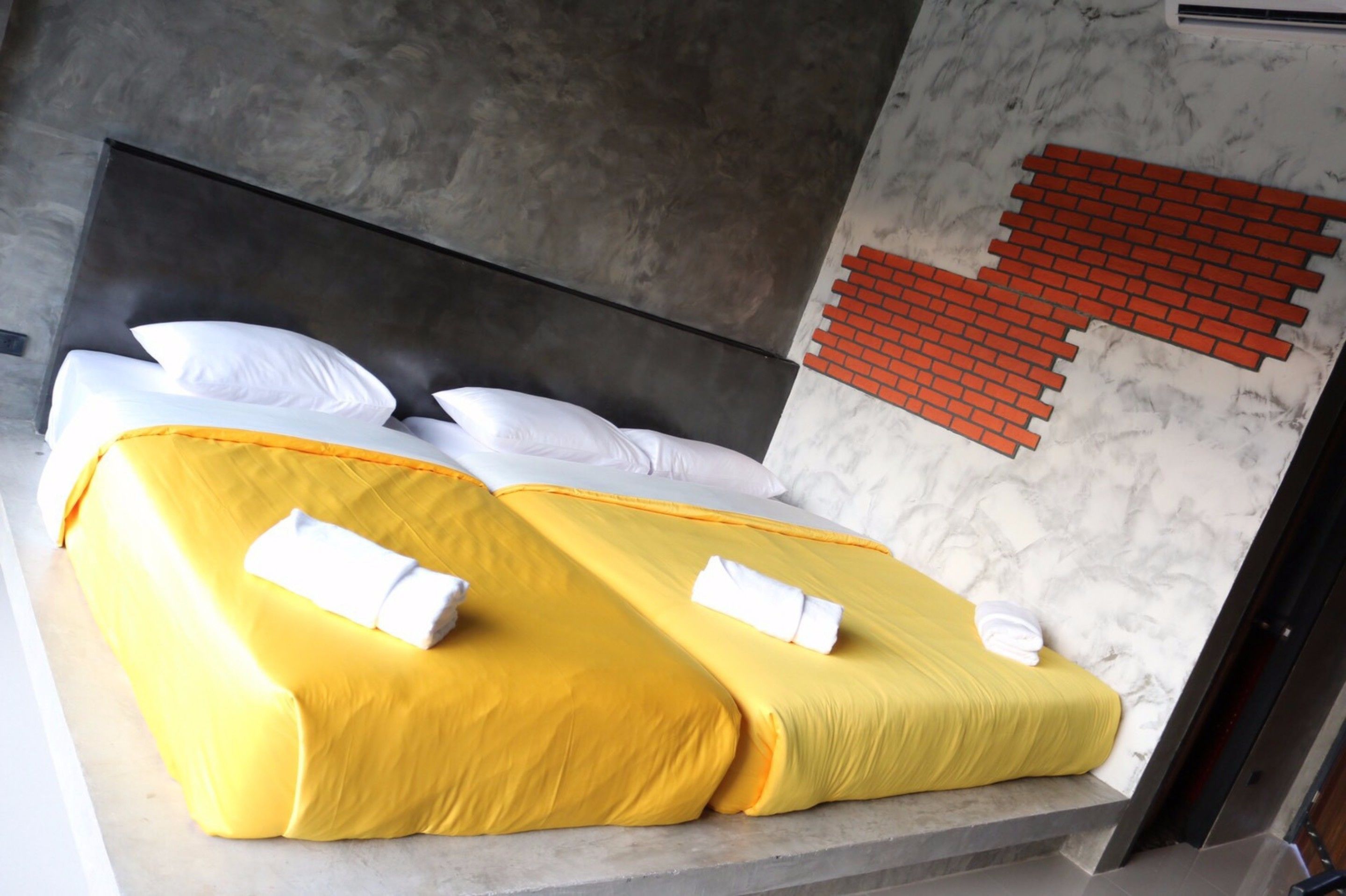 A double bed with yellow bedding and white pillows, a dark headboard, and a red brick pattern on the wall.