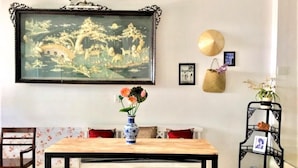 A dining area with a wooden table, chairs, a vase with flowers, a framed painting, a decorative basket, and a small shelf with a picture. - Rubis Hostel (Hue)