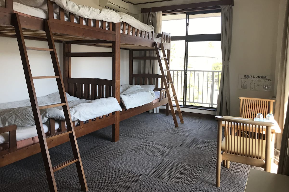 Una habitación con literas, una escalera de madera, una silla y un ventanal con cortinas.