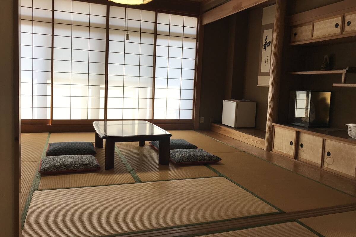 Una habitación japonesa tradicional con suelo de tatami, una mesa baja, cojines, puertas correderas shoji y un televisor.