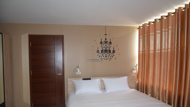 A hotel room with a wooden door, a bed with white bedding, a chandelier, and brown curtains.