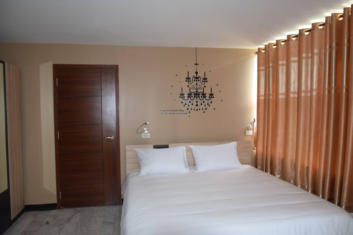 A hotel room with a wooden door, a bed with white bedding, a chandelier, and brown curtains.