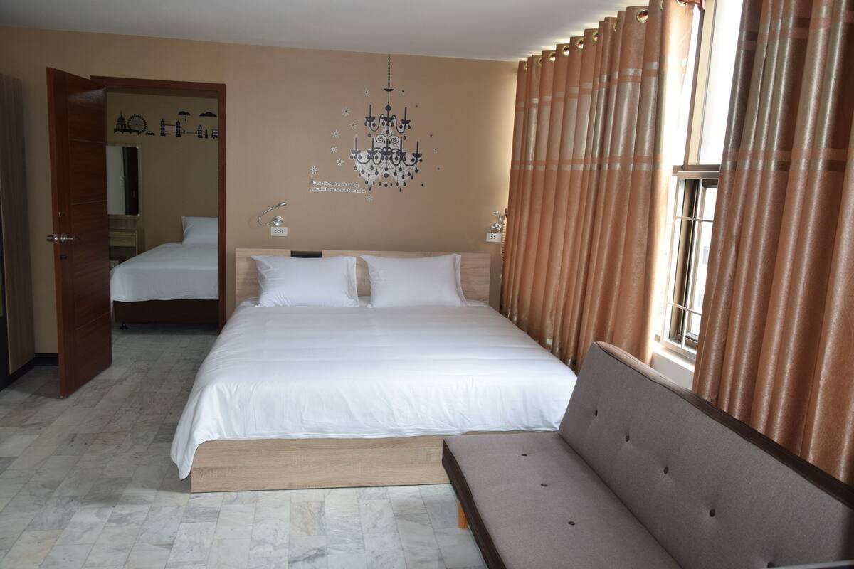 A hotel room with a large bed, a bench, a chandelier, and a window with curtains.
