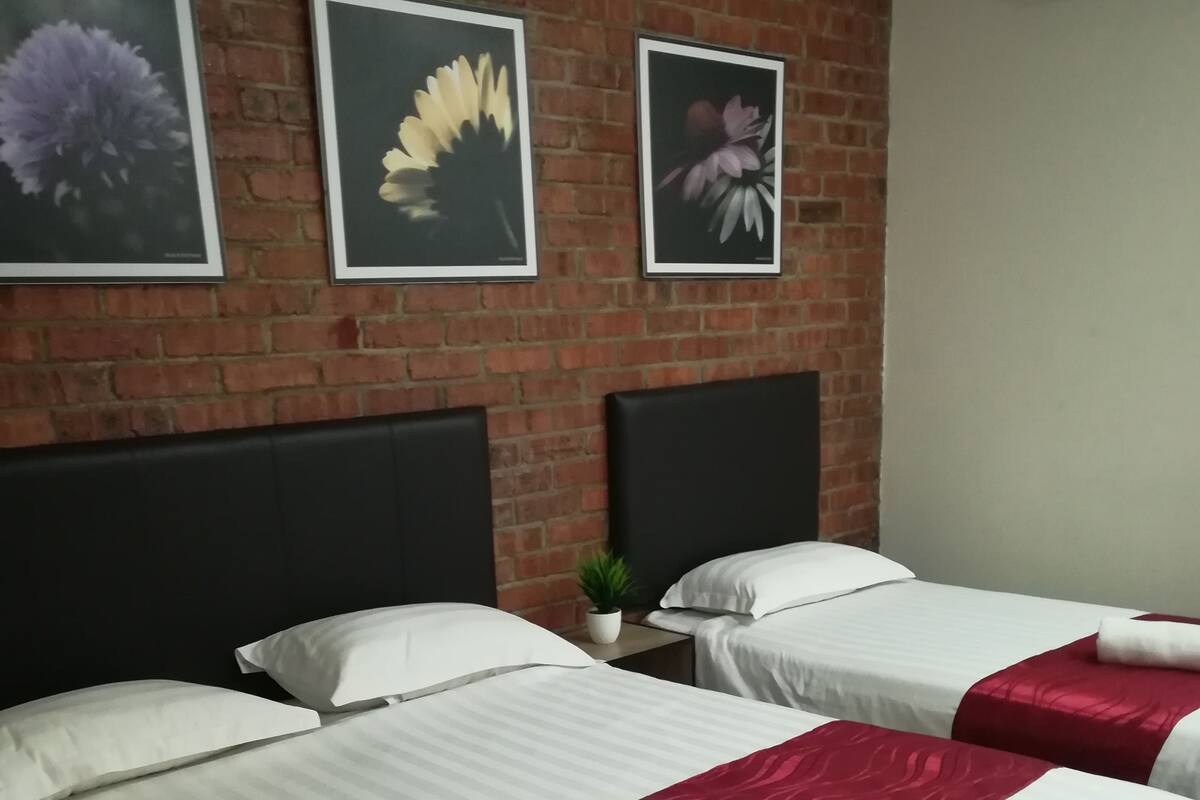A hotel room with two beds, each with white linens and red accents, a brick wall, and framed floral artwork above the beds.