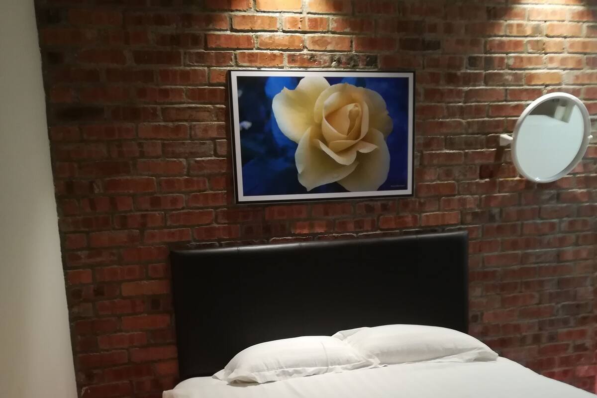 A hotel room with a brick wall, a framed picture of a flower, a round mirror, and a neatly made bed with rolled towels.
