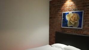 A neatly made bed with white linens and pillows, a brick wall with a framed picture, and a red under-bed lighting strip. - Rock & Roll Hotel (Klang)