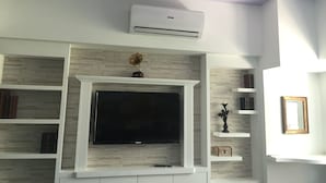 A living room with a flat-screen TV mounted on a stone-textured wall, white shelves with books, a decorative gramophone, and a ceiling-mounted air conditioner. - Kaohsiung Rouen (Kaohsiung)