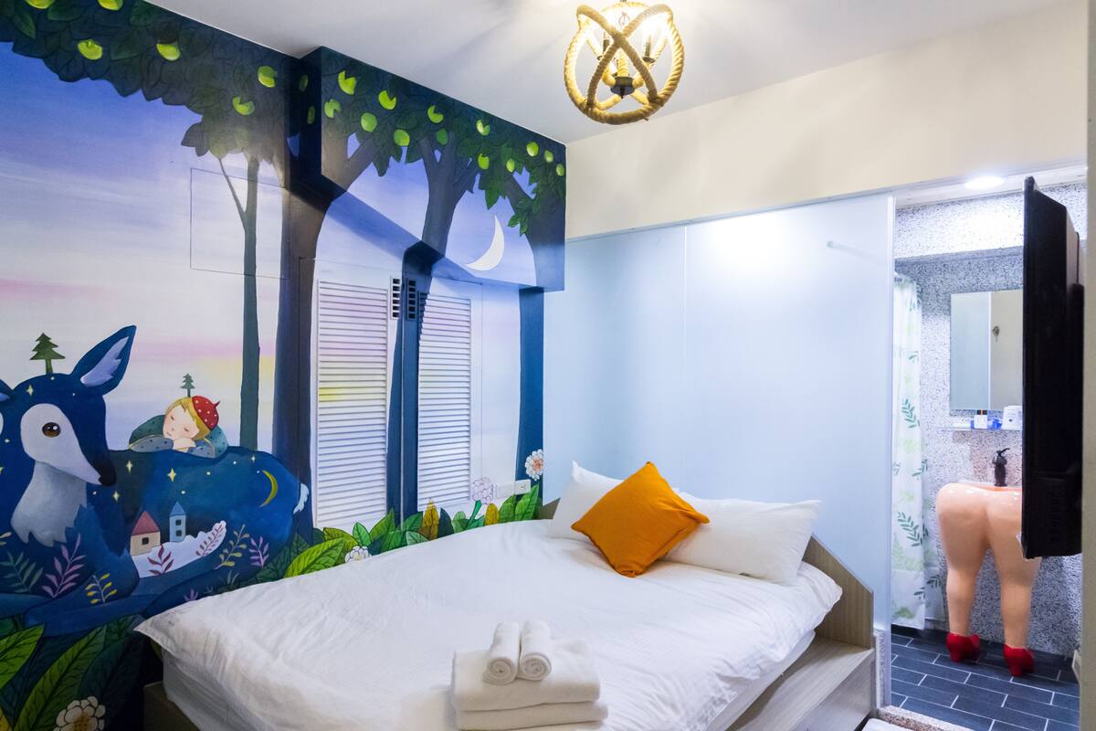 Quarto com cama, painel de parede com céu noturno, janela com persianas e banheiro visível por uma porta aberta.