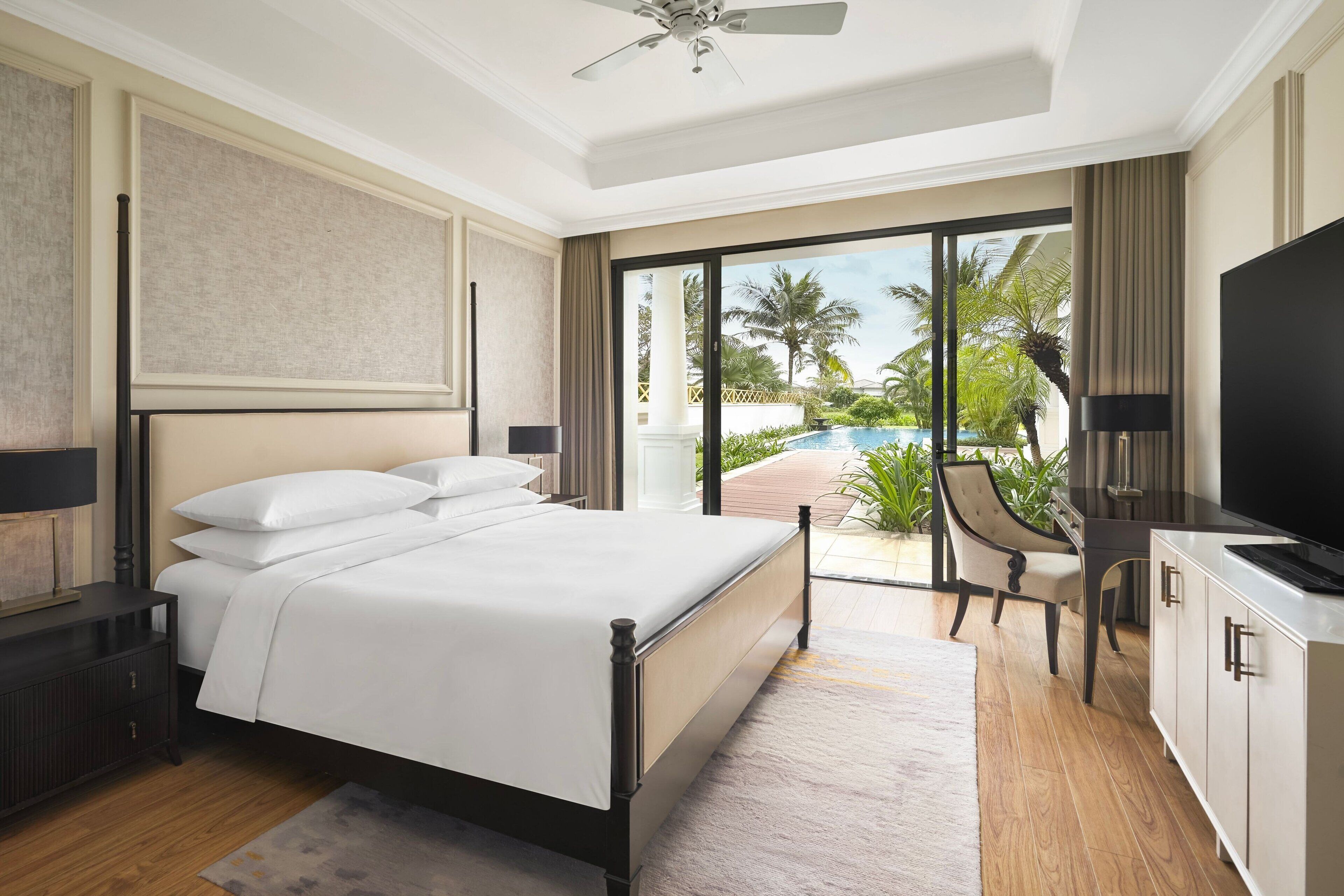 A modern hotel room with a large bed, a TV, a desk, and a view of a swimming pool.