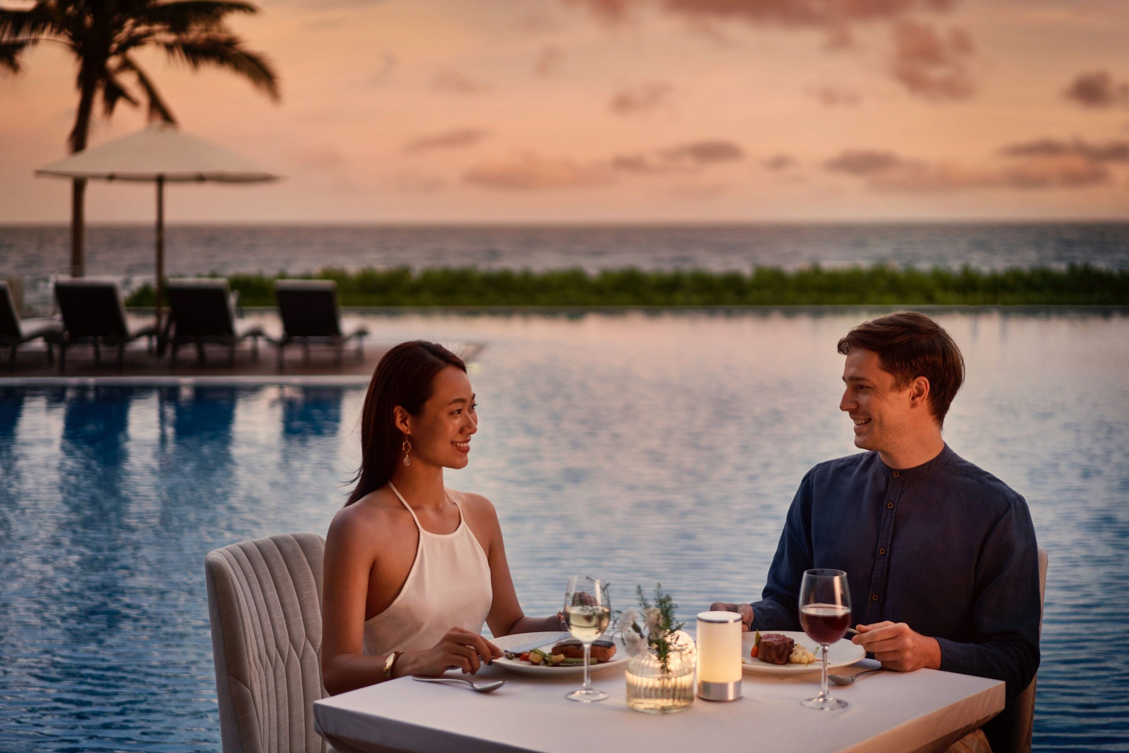 Una pareja cenando junto a una piscina con vista al atardecer, una mesa con bebidas y una vela, y sillones de descanso al fondo.