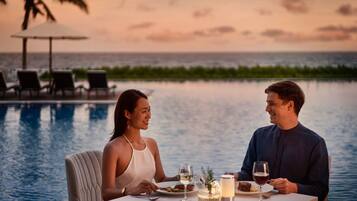 Una pareja cenando junto a una piscina con vista al atardecer, una mesa con bebidas y una vela, y sillones de descanso al fondo.