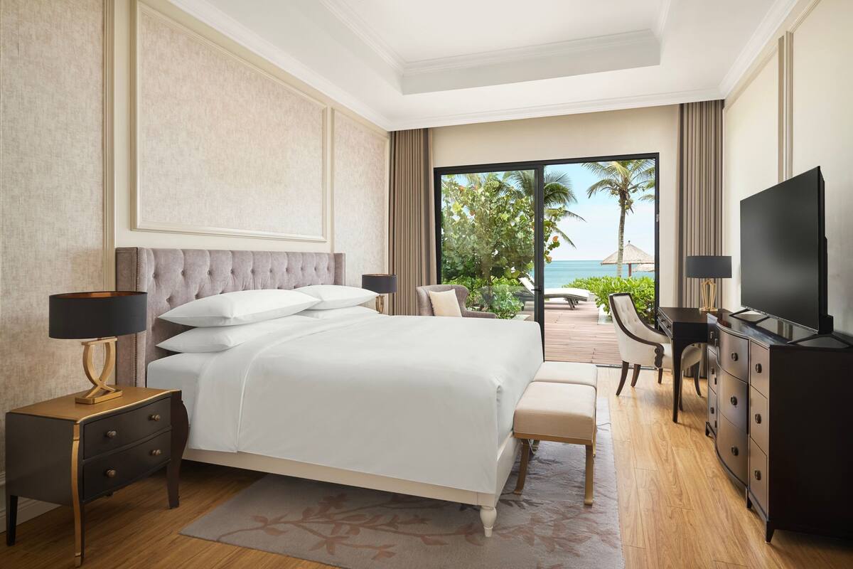 A modern hotel room with a large bed, a desk, a TV, and a balcony with a view of the ocean.