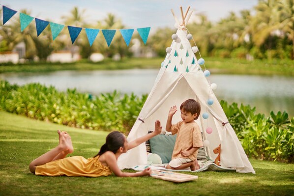 A tent set up on a grassy area near a body of water, with a person lying on the grass and another sitting inside the tent.