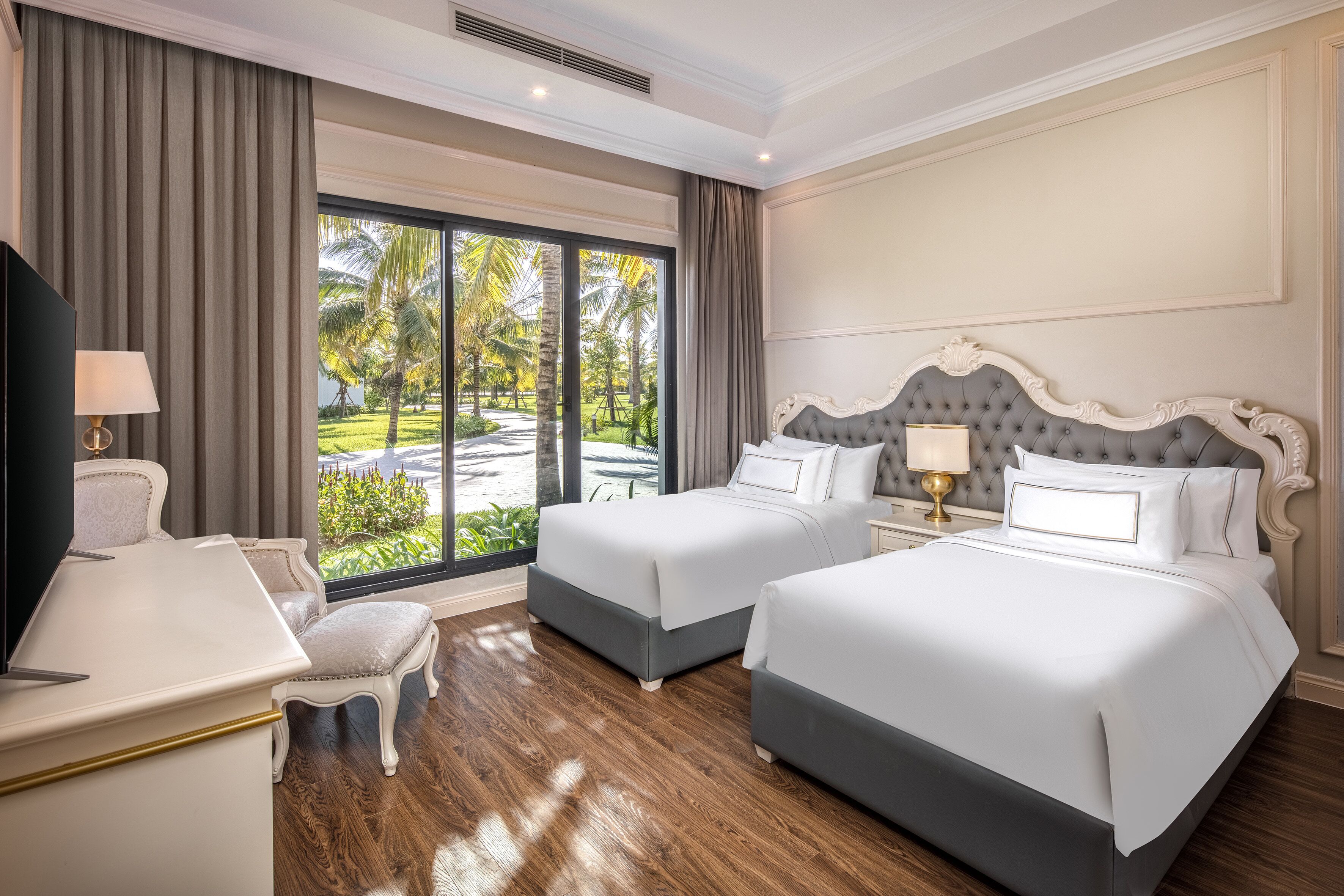 A hotel room with two beds, a desk, and a view of a tropical garden.