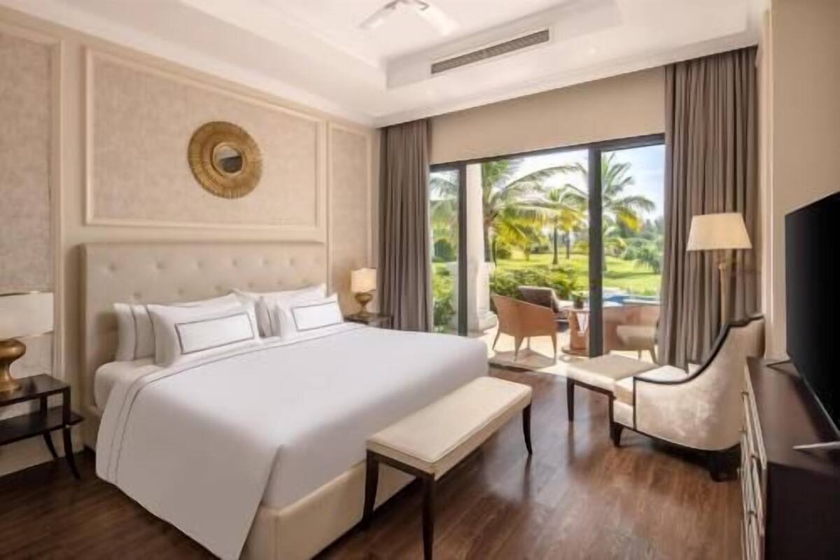 A hotel room with a large bed, a bench, a TV, and a view of a garden.