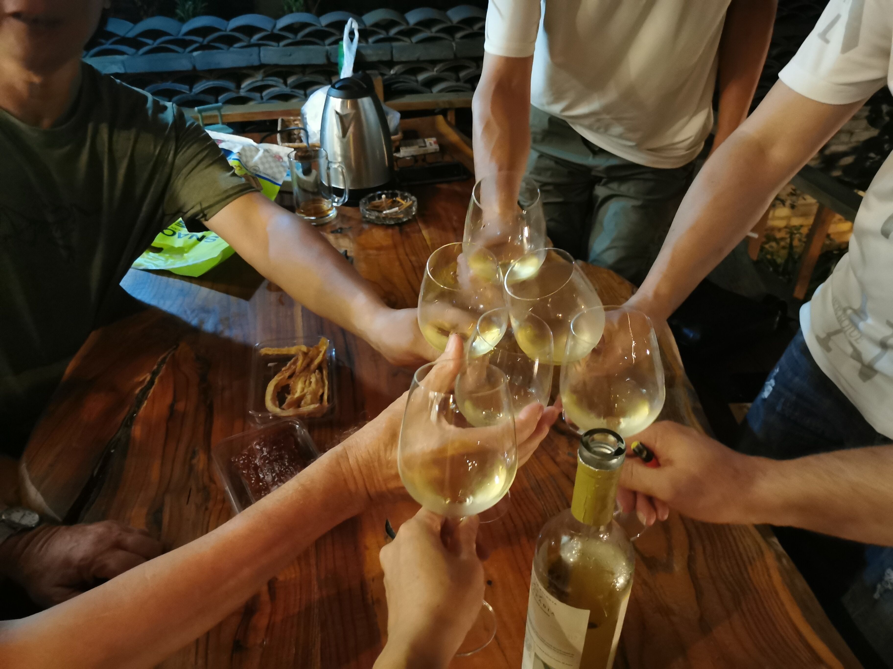 A group of people toasting with wine glasses at a wooden table with a bottle of white wine, a coffee pot, and a small dish of snacks.