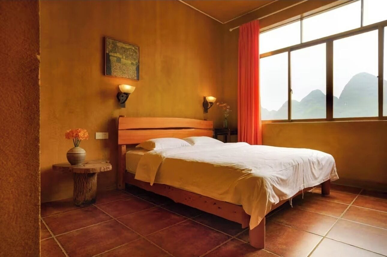 A room with a bed, a window with a view of mountains, a vase with flowers, and wall-mounted lights.