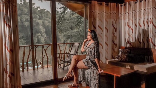 A room with a large sliding glass door leading to a balcony, a wooden floor, a sofa, and a patterned dress. - Kandy Cabana Eco Resort (Weligalla)