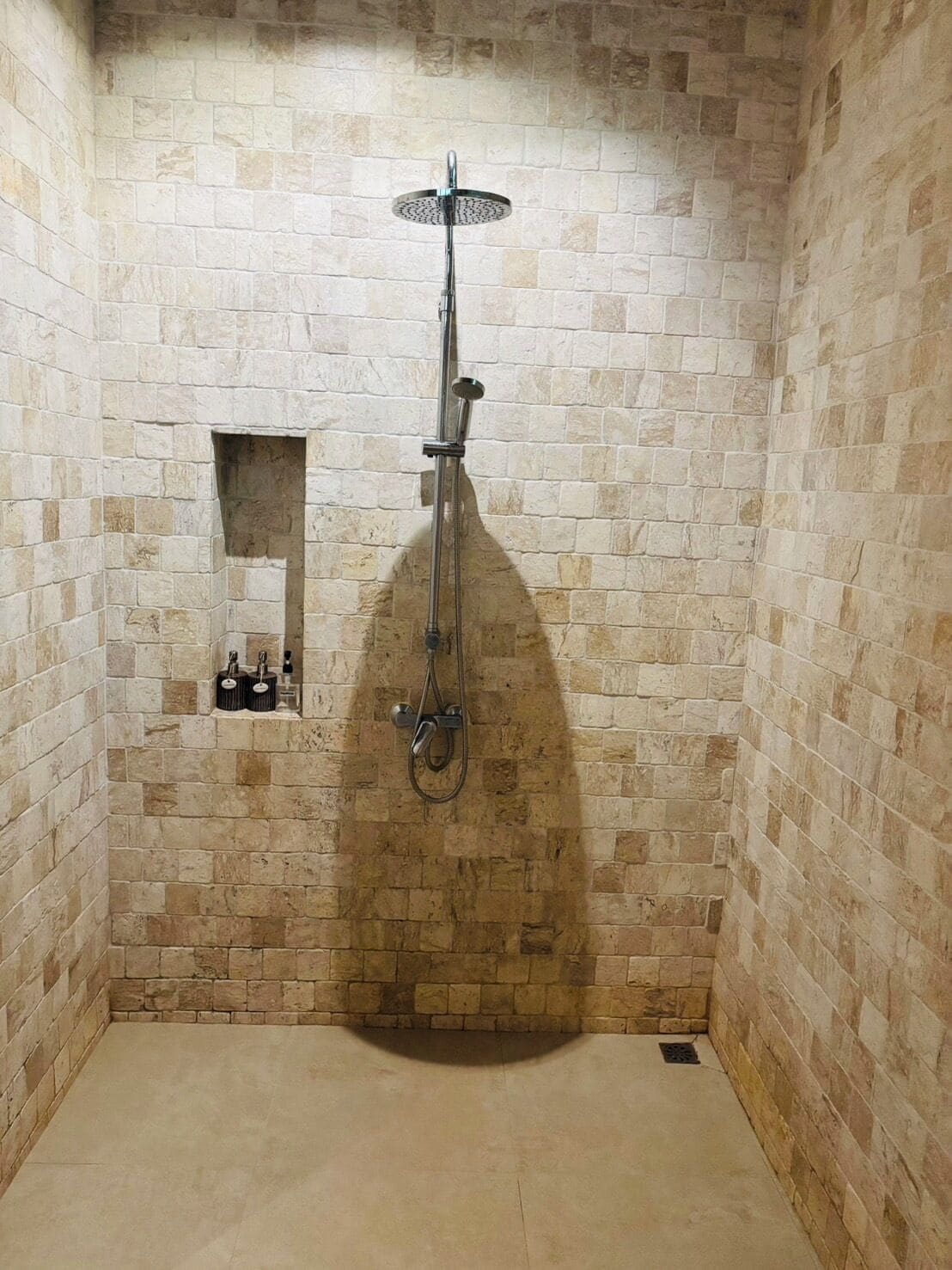 Une douche équipée d’un pommeau de douche à main et d’un robinet mural, installée dans une pièce aux murs en pierre et dotée d’une petite étagère pour les articles de toilette.