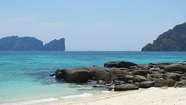 A beach with clear turquoise water, large rocks in the foreground, and distant mountains. - Viking Nature Resort (Ko Phi Phi)