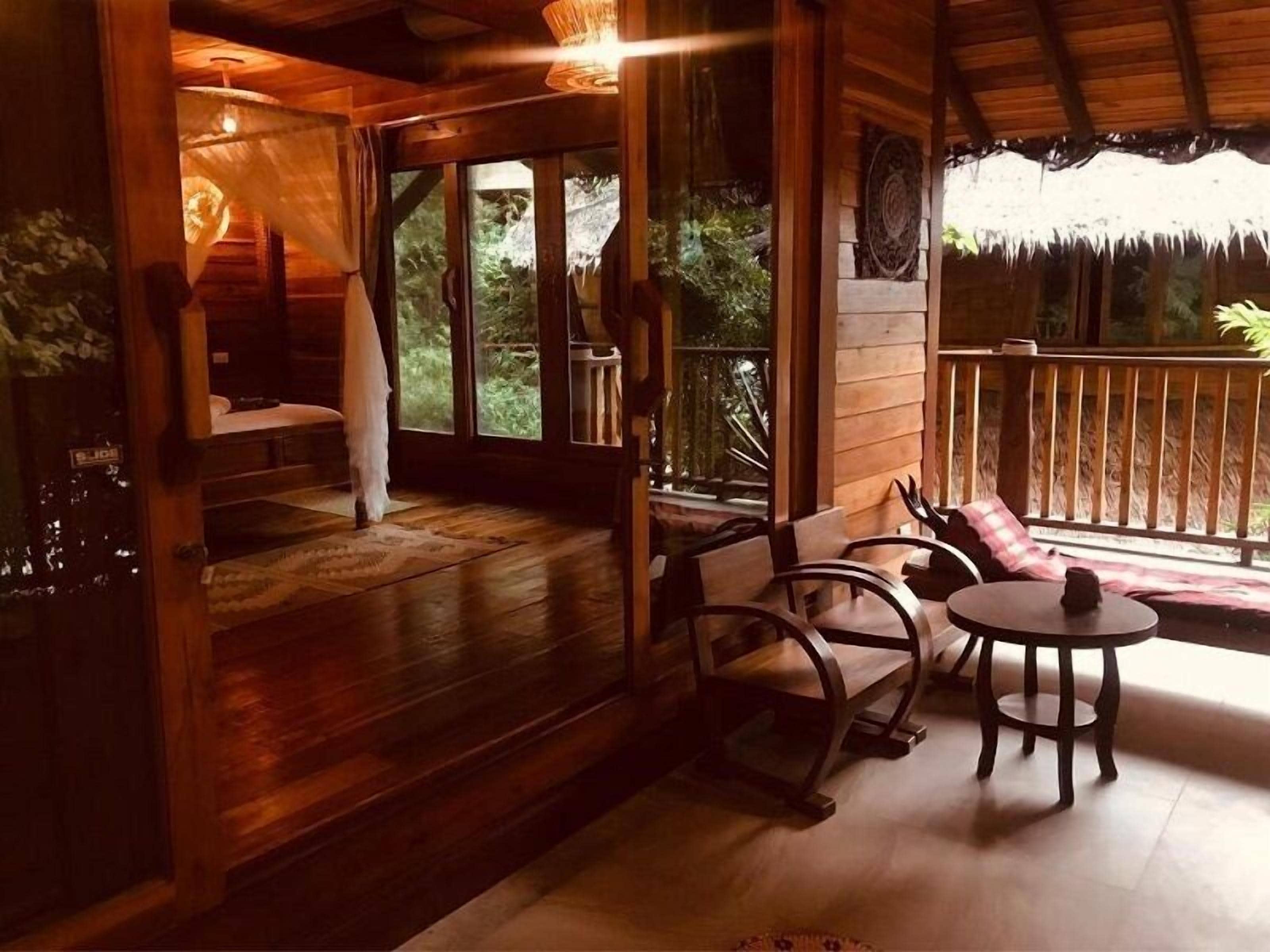 A wooden cabin with a cozy interior, featuring a bed, a seating area with chairs and a table, and large windows offering a view of the surrounding forest.