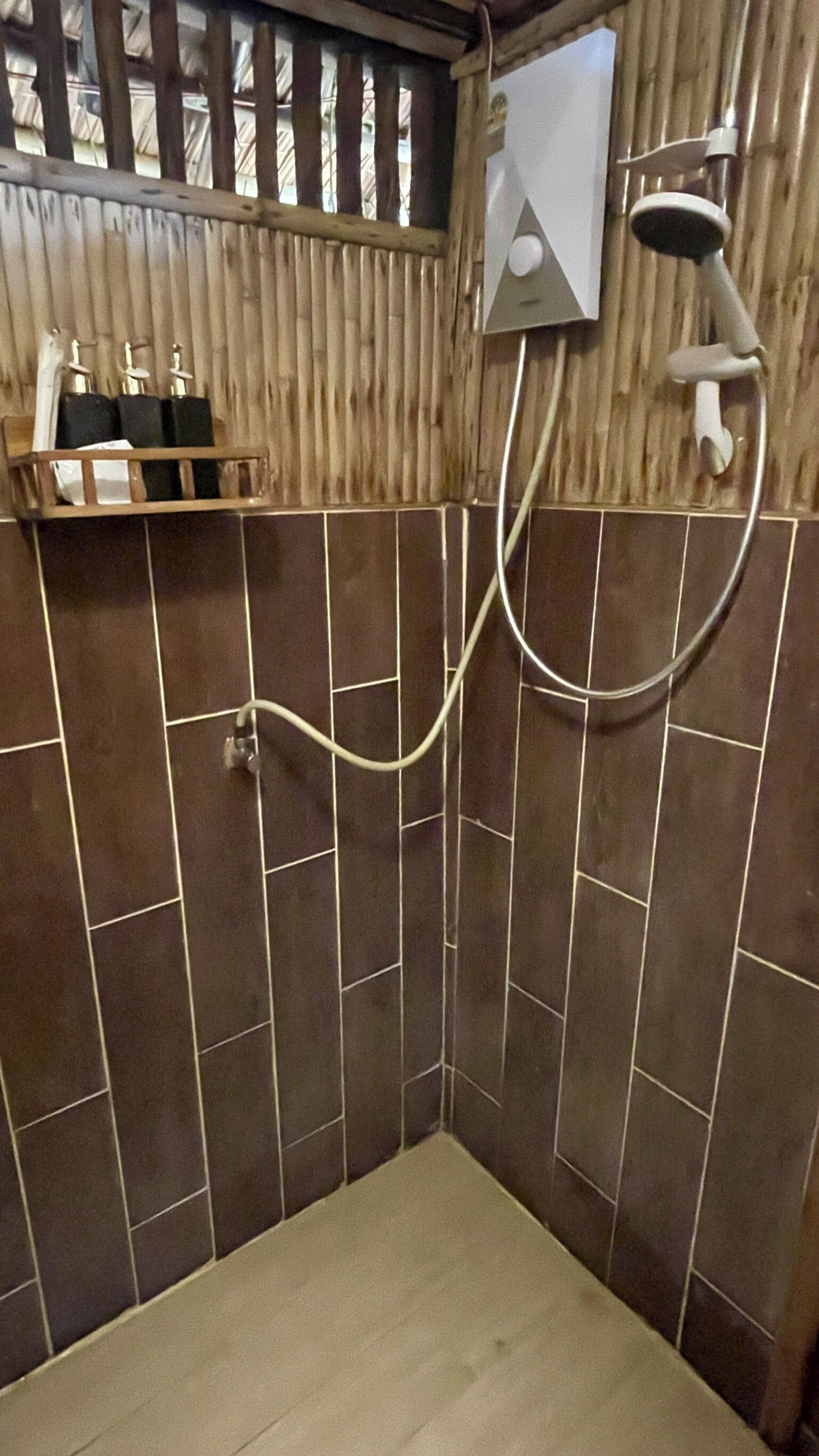A bathroom with dark tiled walls, a shower, and a wooden shelf with toiletries.