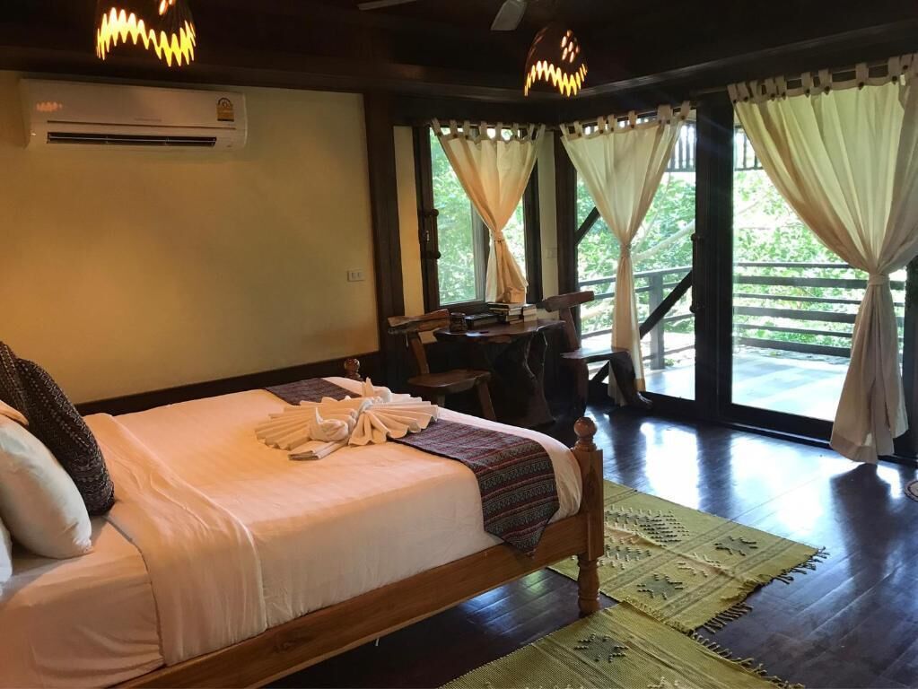 A neatly made bed with a blanket and pillows, a wooden desk with a chair, and a view of greenery through large windows.
