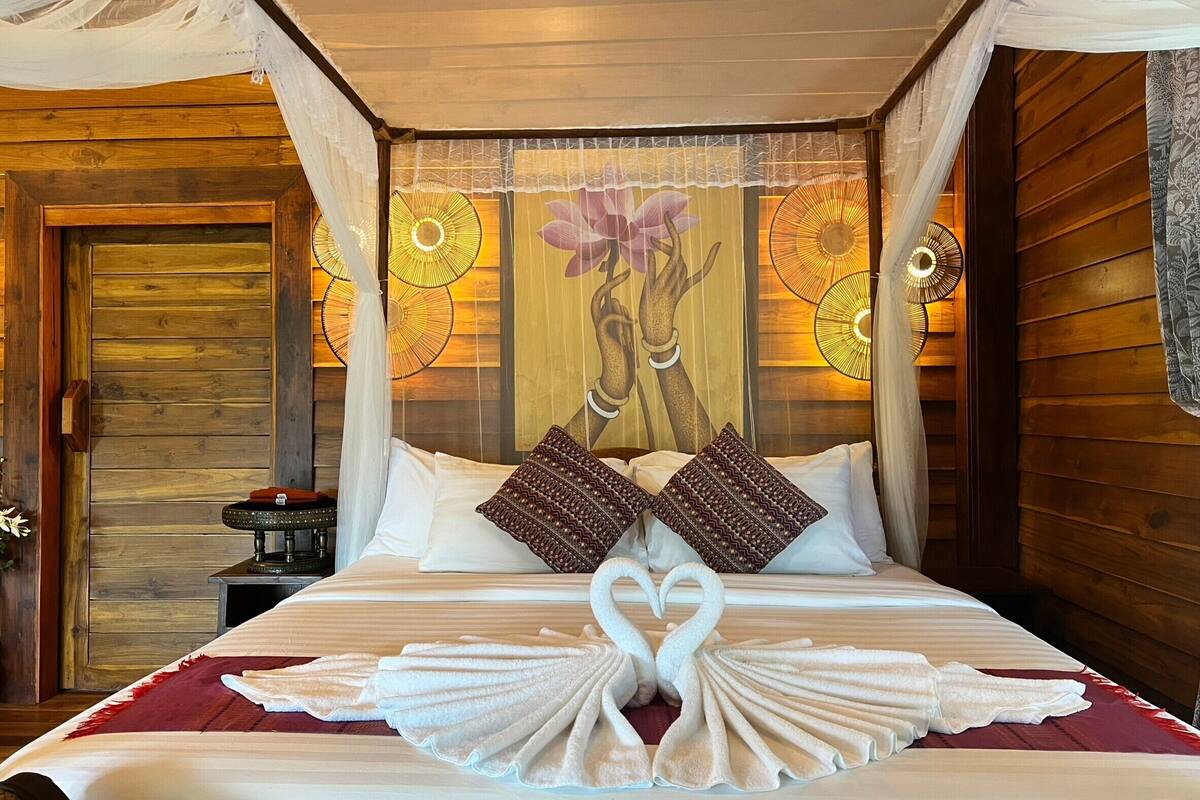 En pænt redt seng med et hjerteformet håndklædearrangement, et træsengbord og en væg med et blomstermural.
