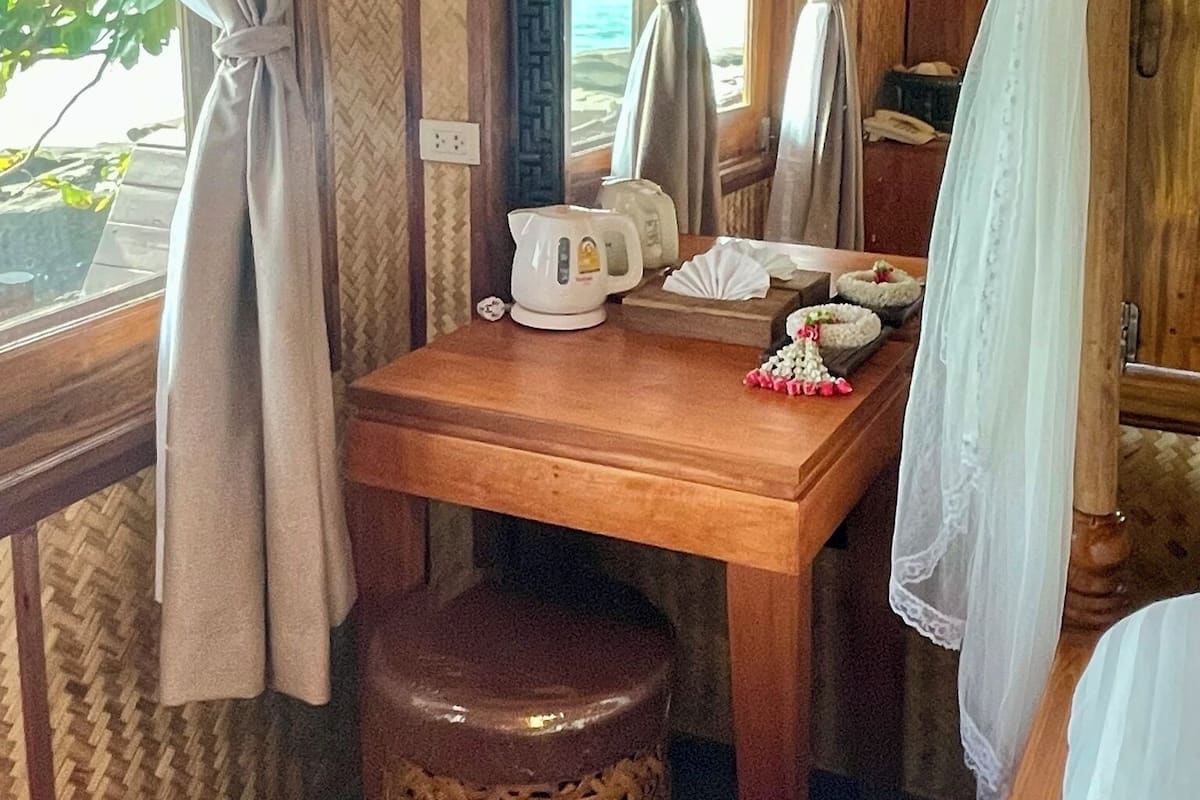 A room with a wooden table, a white kettle, a small stool, and a view of the sea through curtains.