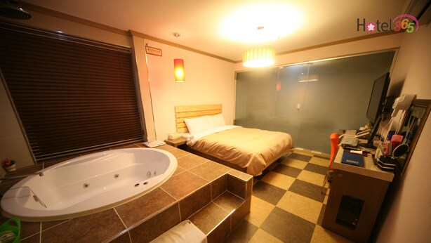 A hotel room with a large bathtub, a bed with a wooden headboard, a checkered floor, and a small desk with a mirror and toiletries. - Chuncheon Myeongdong Hotel (Chuncheon)