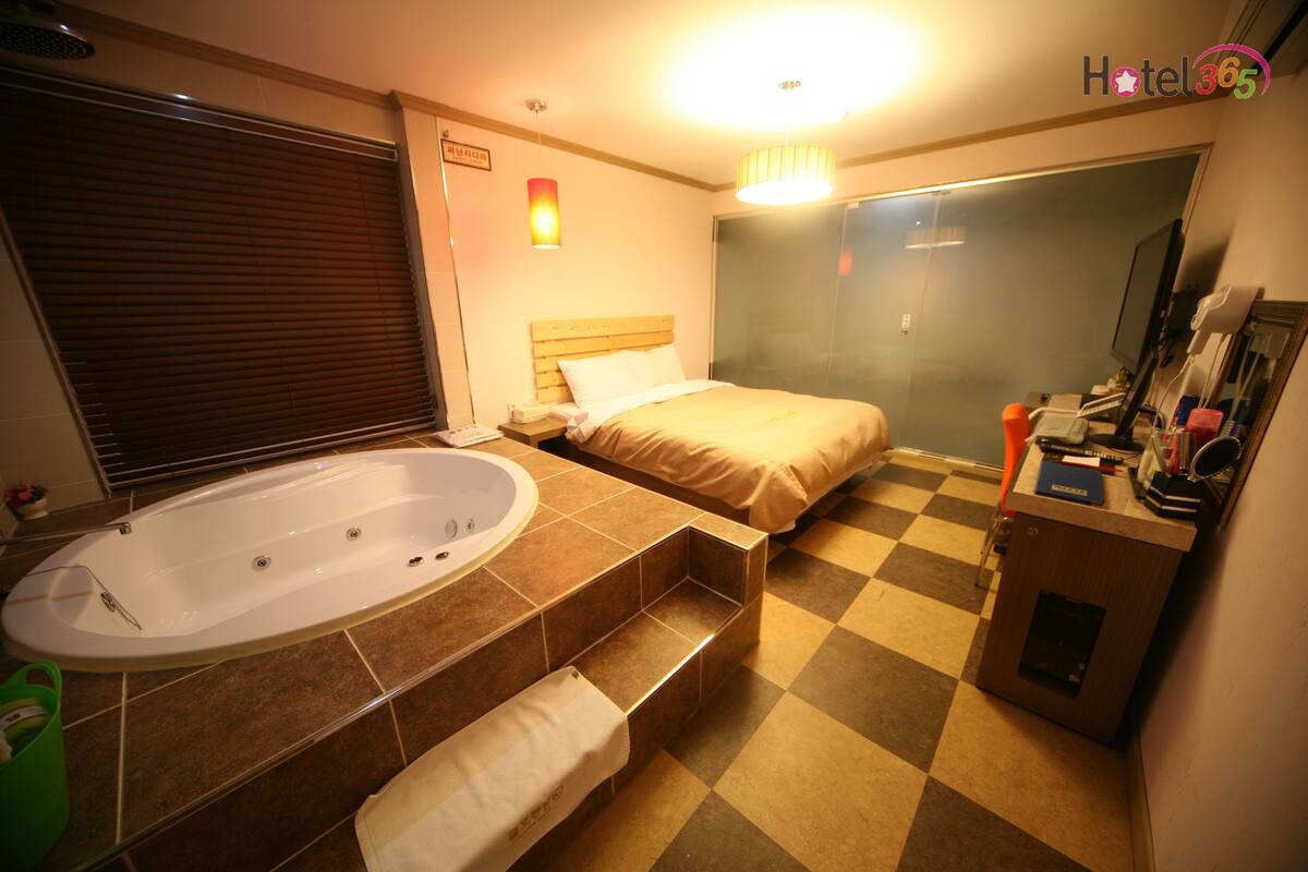 Habitación de hotel con una bañera grande, una cama con cabecera de madera, suelo a cuadros y un escritorio pequeño con un espejo y artículos de aseo.