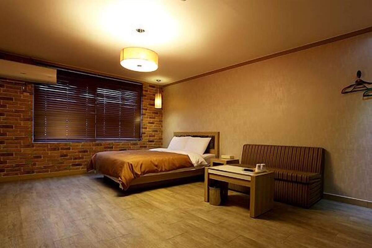 Habitación de hotel con cama, una mesita, una silla, ventana con persianas y pared de ladrillo.