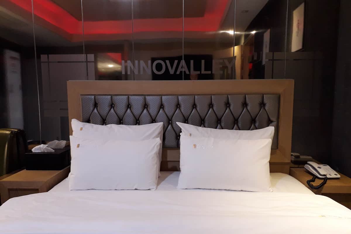 A hotel room with a tufted headboard, two white pillows, a neatly made bed, a bedside table with a phone, and a sign reading "INNOVALLEY".