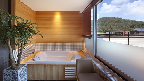 Un baño moderno con pared de madera, una bañera grande, una zona de estar y vistas a un edificio y colinas.
