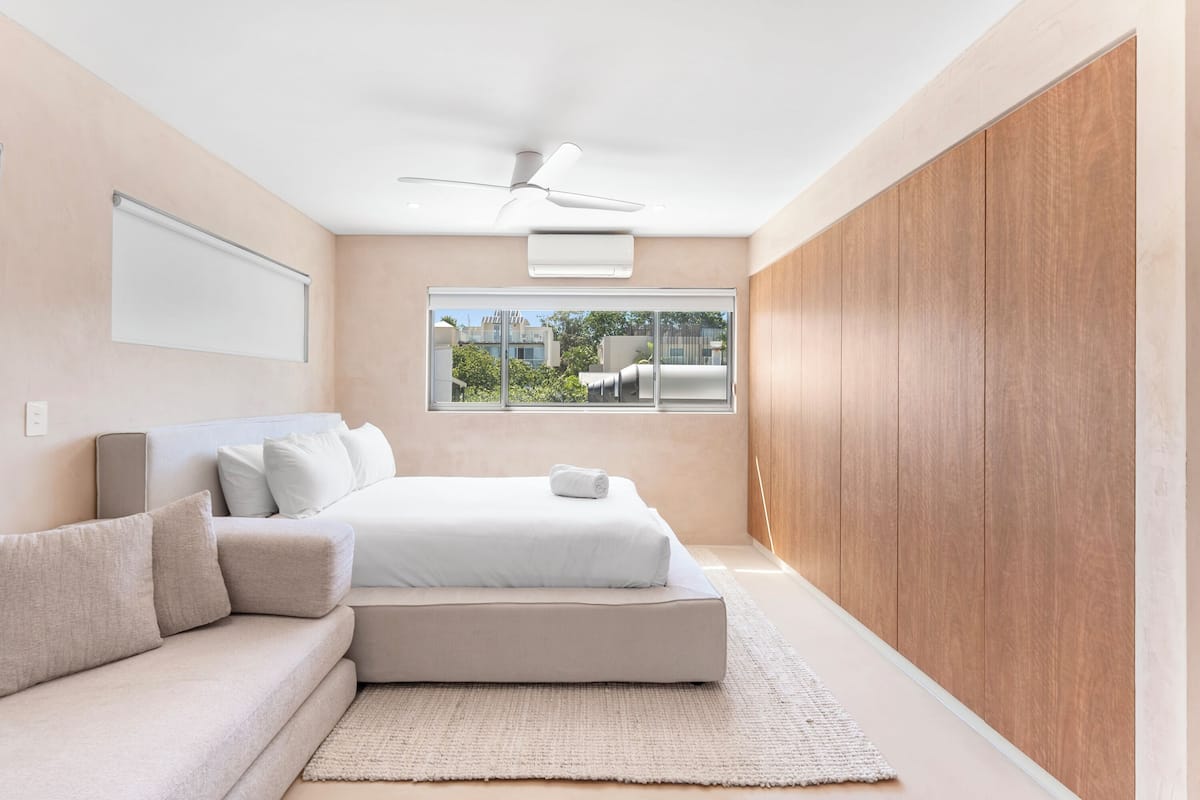Ein modernes Schlafzimmer mit Bett, Sofa, Deckenventilator und einem Fenster mit Blick auf Gebäude.