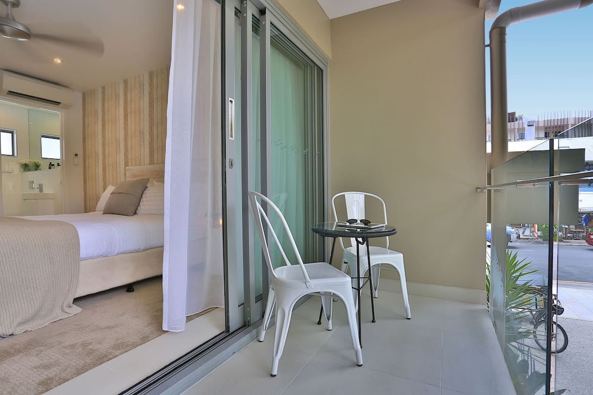 Ein Schlafzimmer mit Bett, einer Glastür, die auf einen Balkon führt, einem Sessel und einem kleinen Tisch.
