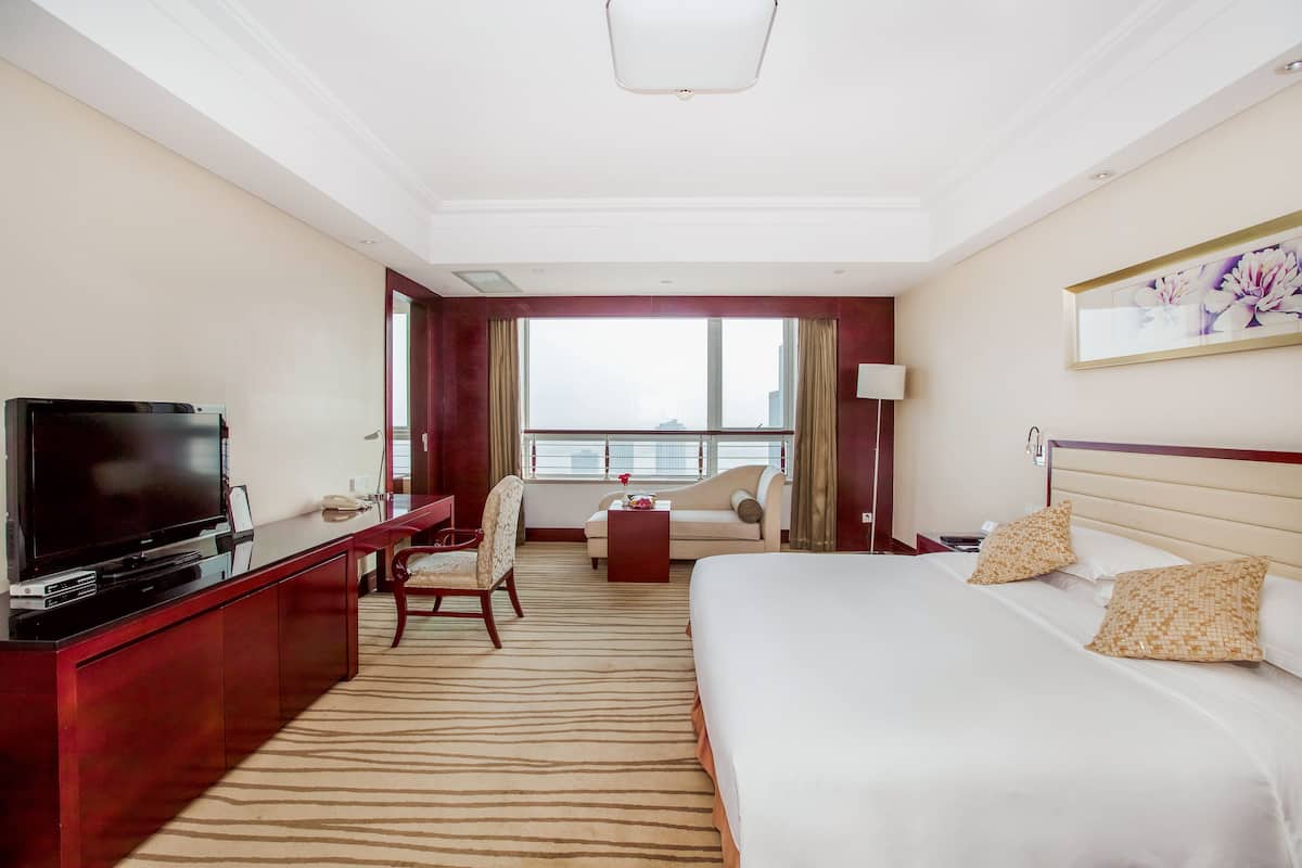 Habitación de hotel con una cama grande, un escritorio con televisor, un sofá y vistas a la ciudad a través de amplios ventanales.