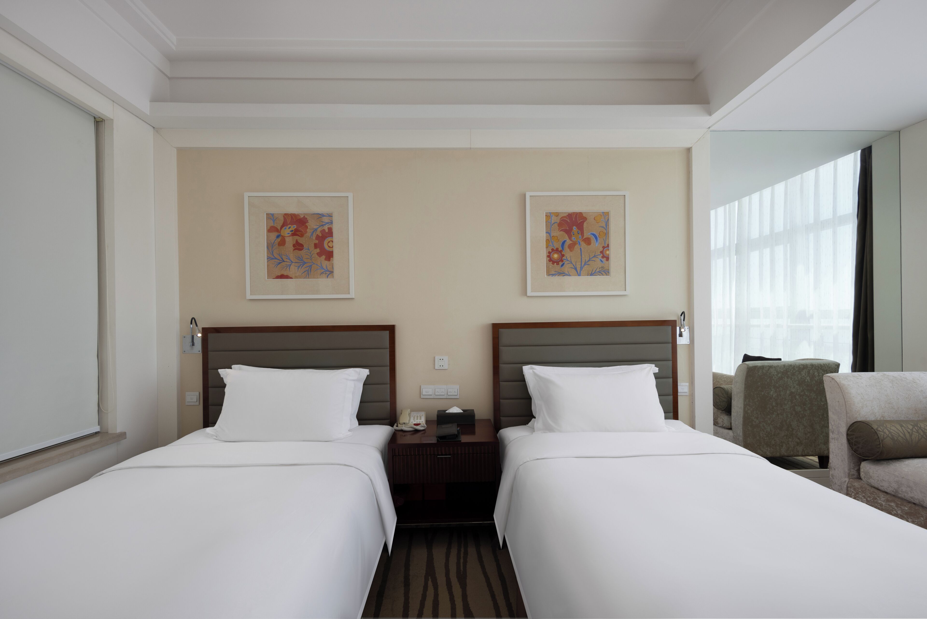 Habitación de hotel con dos camas, una mesita de noche, una silla y dos cuadros con motivos florales en la pared.