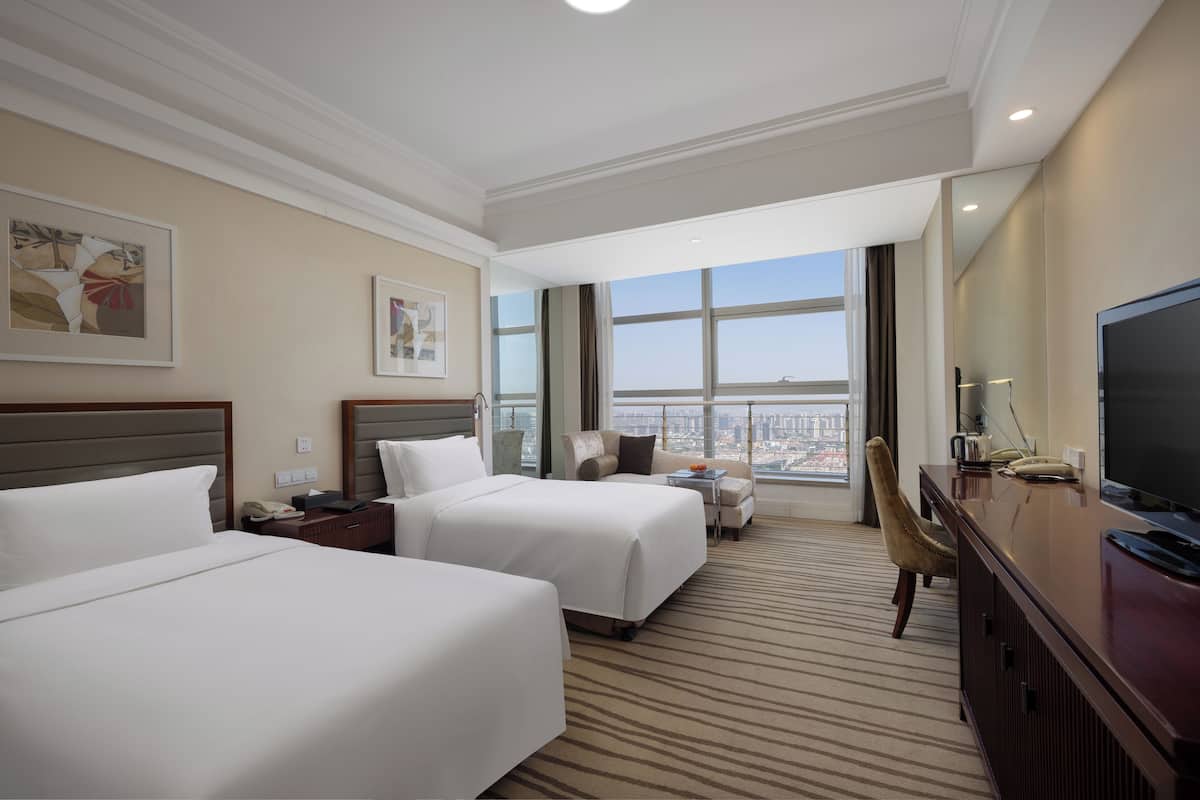 Habitación de hotel con dos camas, un escritorio, una silla, un televisor y ventanas amplias con vistas a la ciudad.