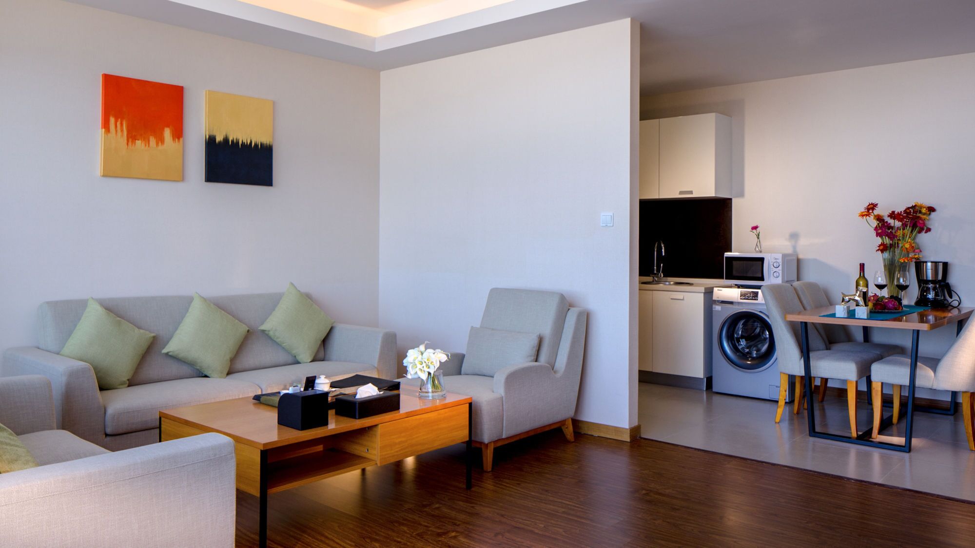 A modern living room with a sofa, armchairs, a coffee table, and a dining area with a table and chairs. A kitchen with a washing machine and a microwave is visible in the background.