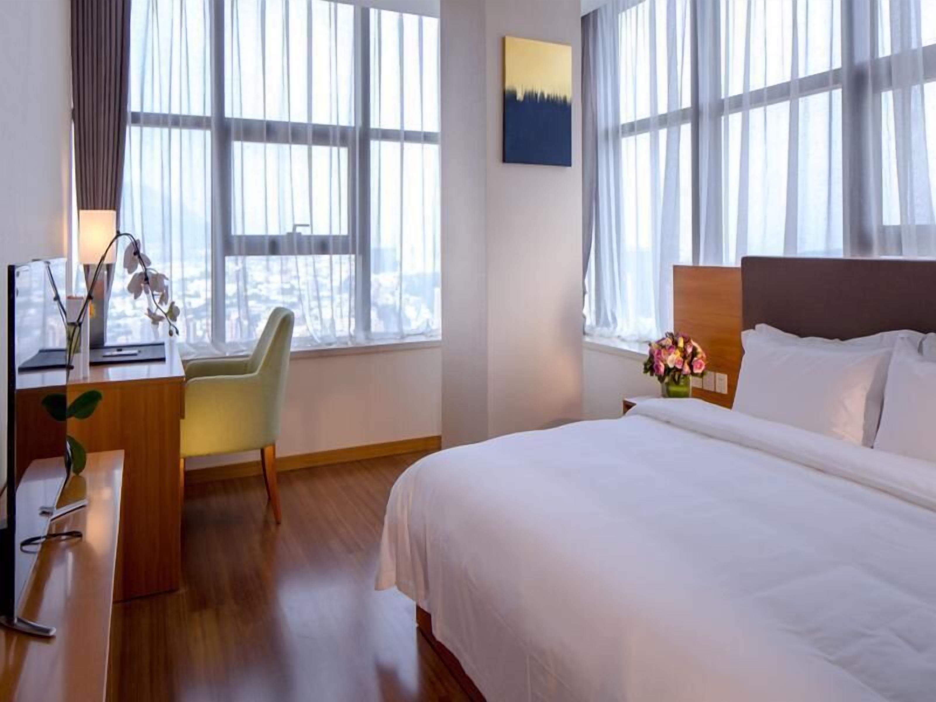 A hotel room with a large bed, a desk with a lamp, a chair, and a view of the city through large windows.