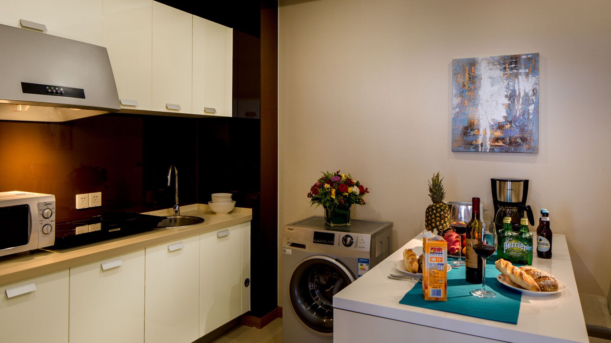 A compact kitchen with a microwave, sink, and washing machine. A painting hangs on the wall, and a table with food and drinks is visible.