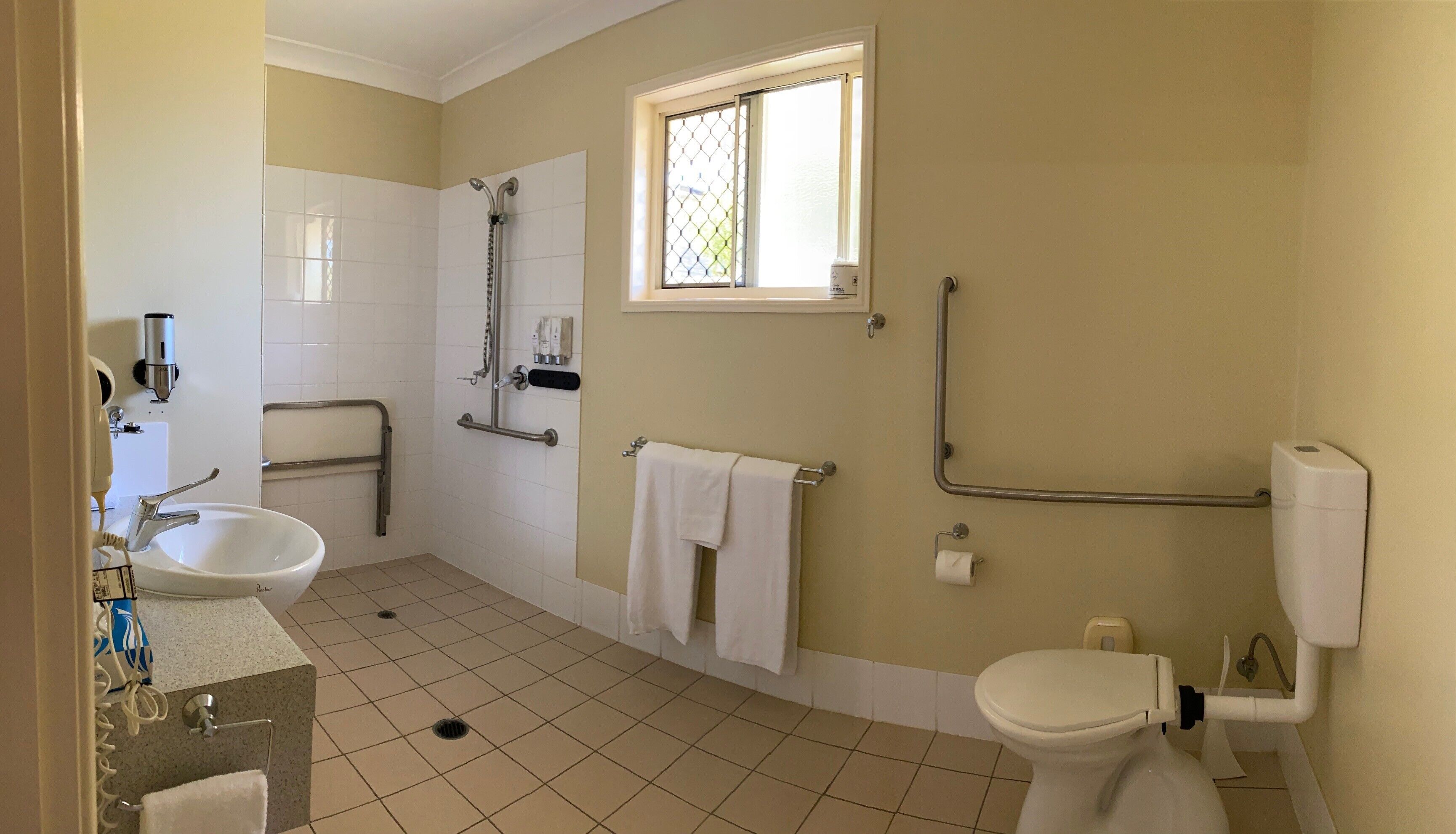 Un bagno con lavandino, toilette e doccia. Una finestra permette l'ingresso di luce naturale, ed è presente una maniglia per facilitare l'accesso.