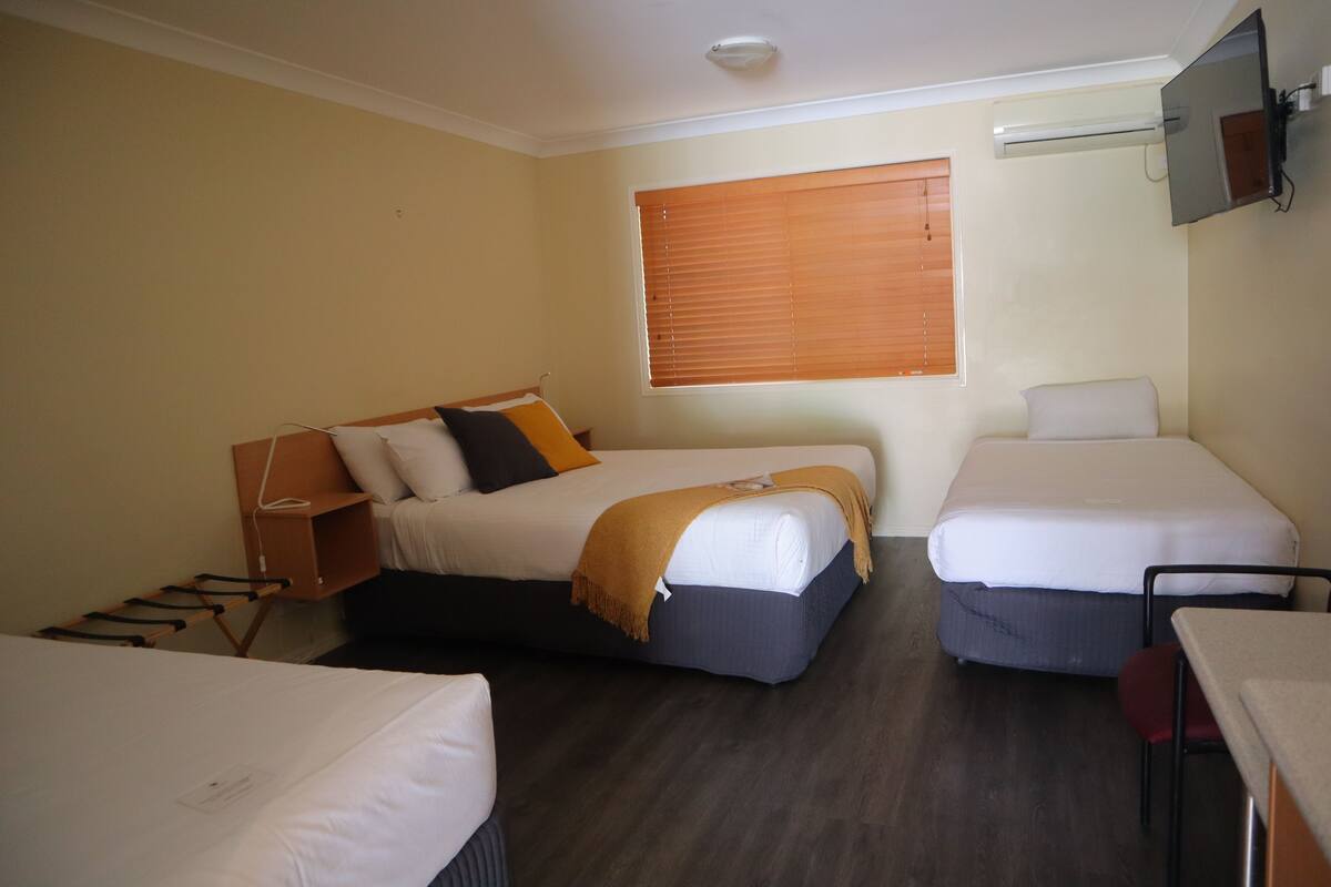 A hotel room with two single beds, a desk, a chair, a TV mounted on the wall, and a window with blinds.